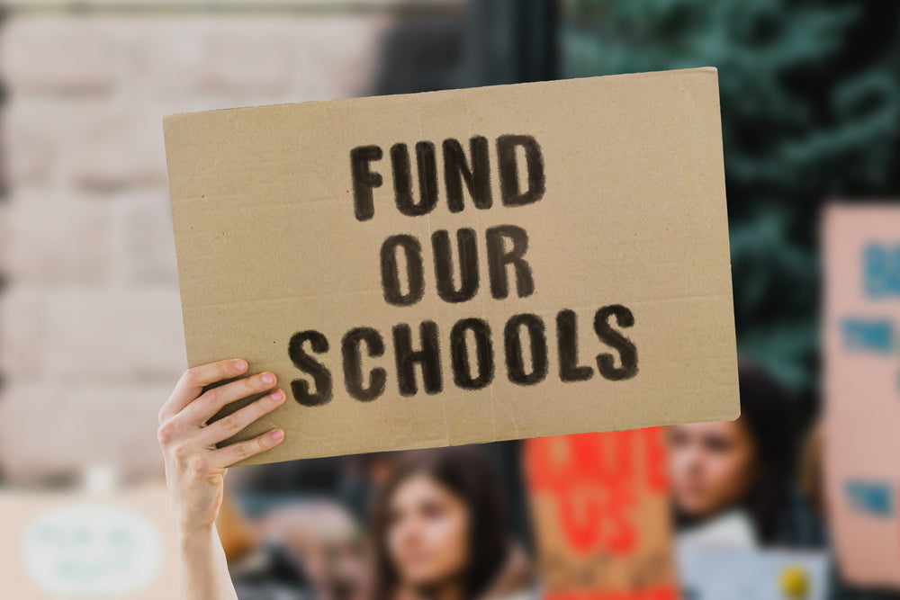 The phrase " Fund our schools " on a banner in men's hand. Human holds a cardboard with an inscription